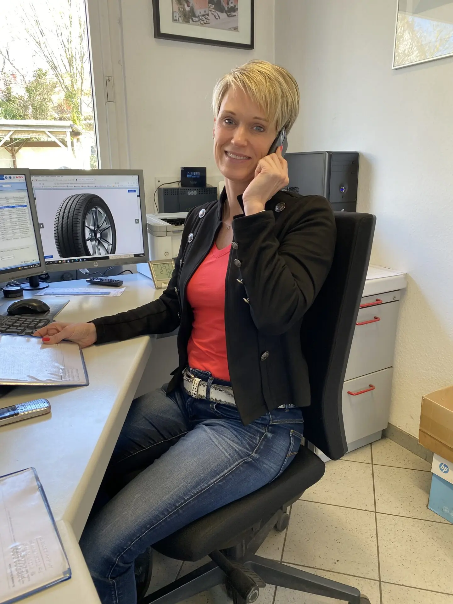 Frau mit blonden Haaren sitzt am Schreibtisch und spricht am Telefon, während sie vor einem Computer sitzt.