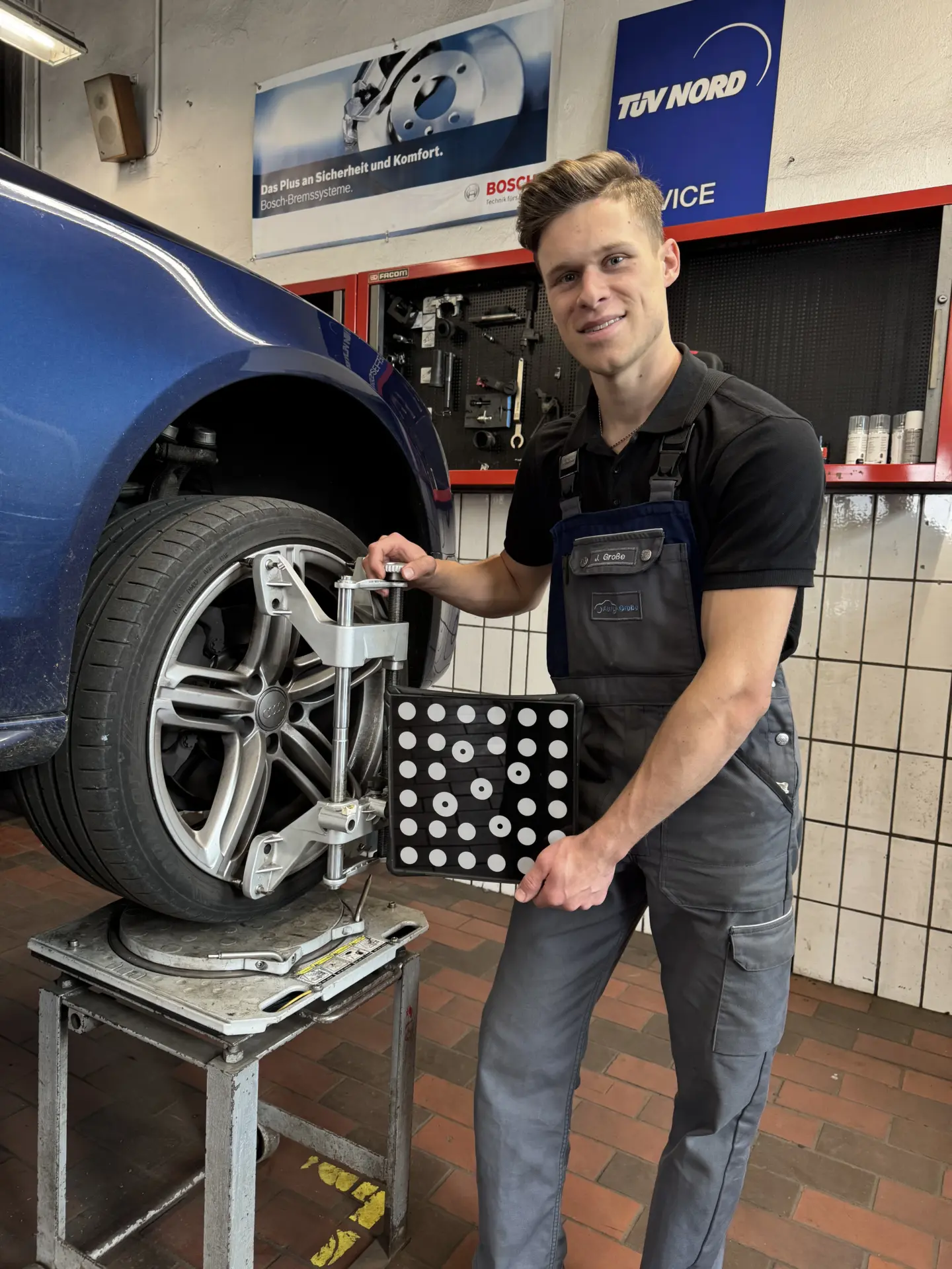 Mechaniker hält ein Achsmessgerät neben einem Auto mit einem abgenommenen Rad in einer Werkstatt.