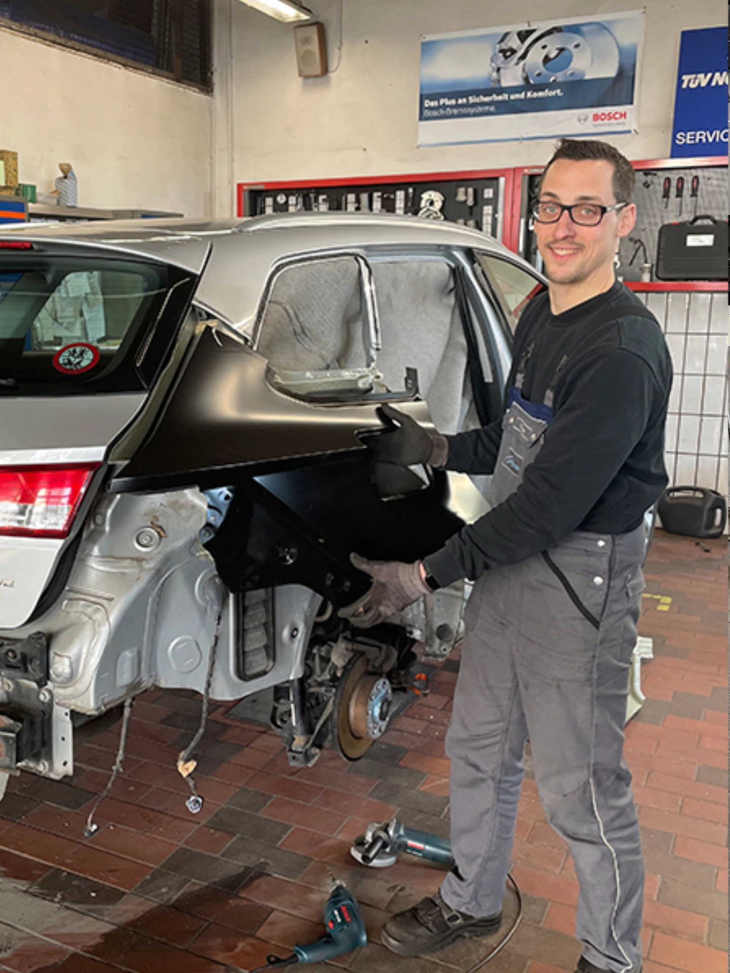 Mechaniker, der ein Fahrzeugteil in einer Werkstatt hält, mit Werkzeugen und Fahrzeugteilen im Hintergrund.