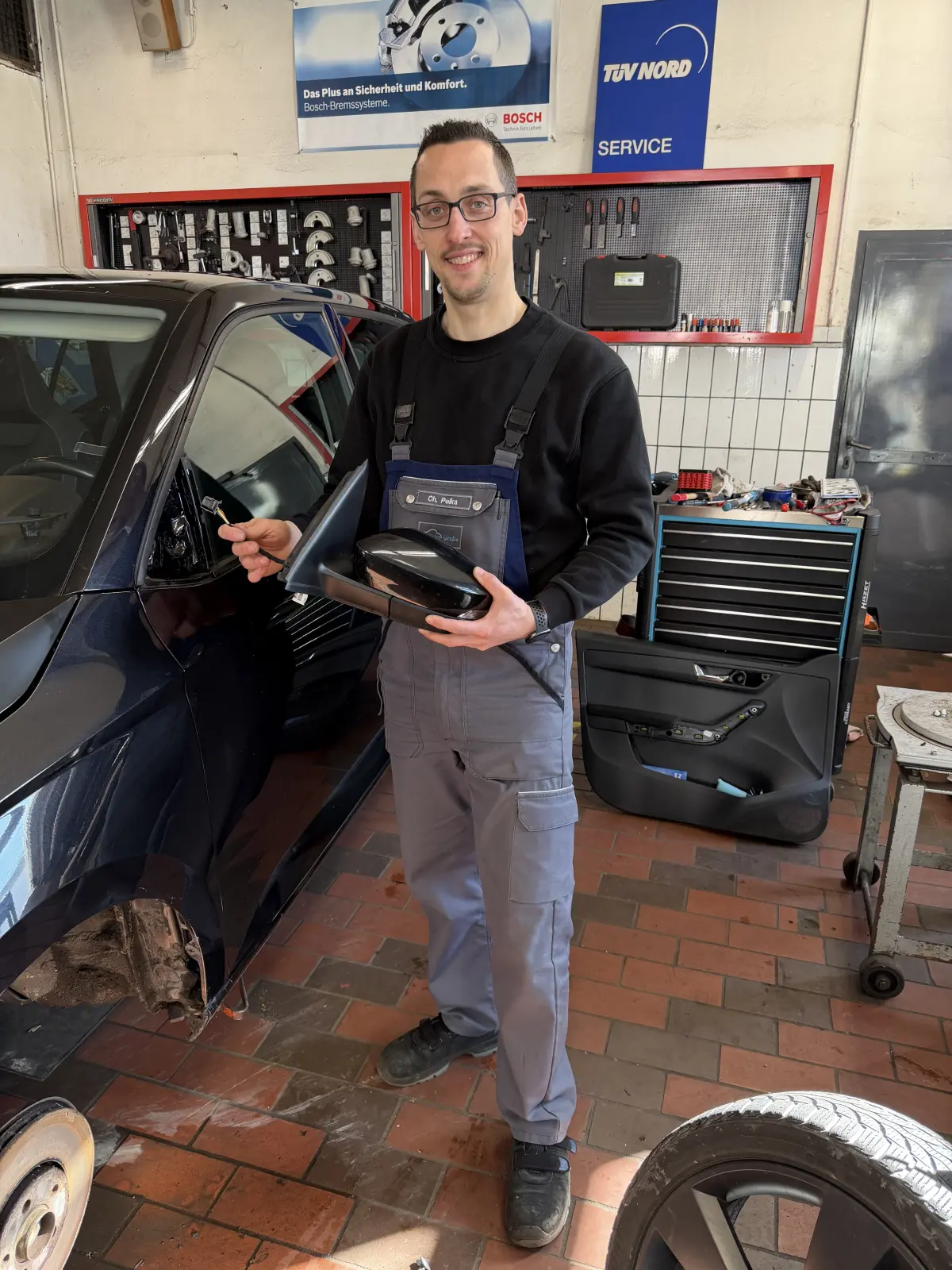 Techniker der KFZ-Reparaturwerkstatt Auto Große hält ein Ersatzteil neben einem schwarzen Auto, in einer Werkstatt mit roter Werkbank und Werkzeuge im Hintergrund.