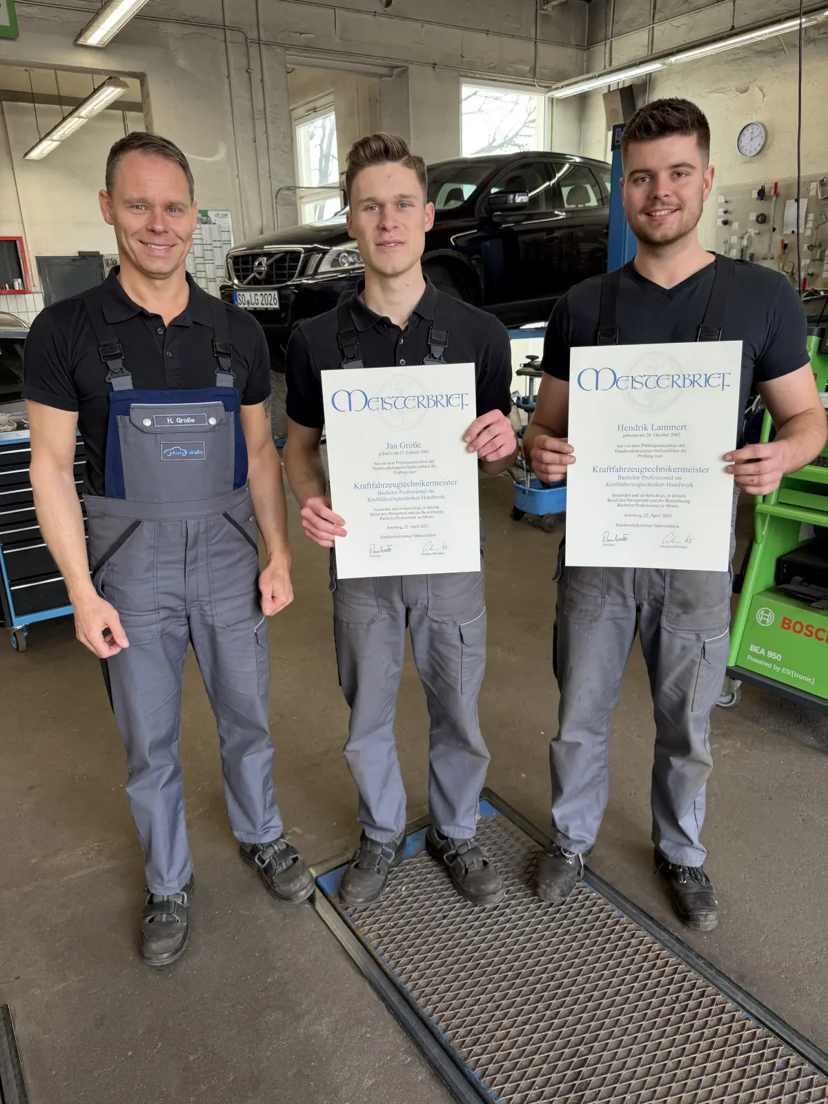 Auto Große Werkstatt: Drei Mechaniker in grauen Hosen und schwarzen Oberteilen halten Zertifikate, im Hintergrund ein Auto auf der Hebebühne in einer Kfz-Werkstatt.