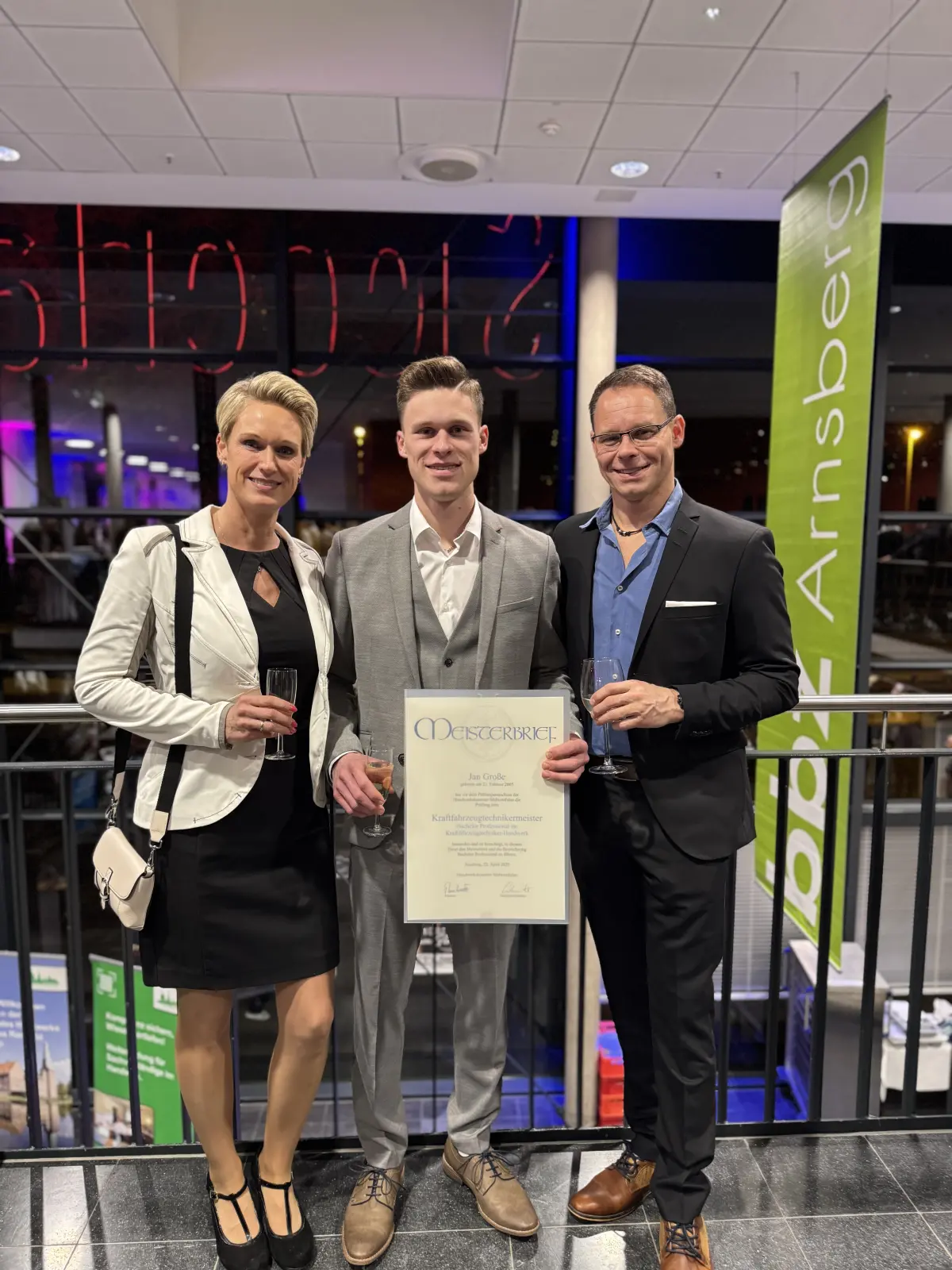 Drei Personen in einem Empfangsbereich einer KFZ-Werkstatt-Veranstaltung, zwei Männer in Anzügen und eine Frau, halten ein Zertifikat; Hintergrundbanner und Glasfront.